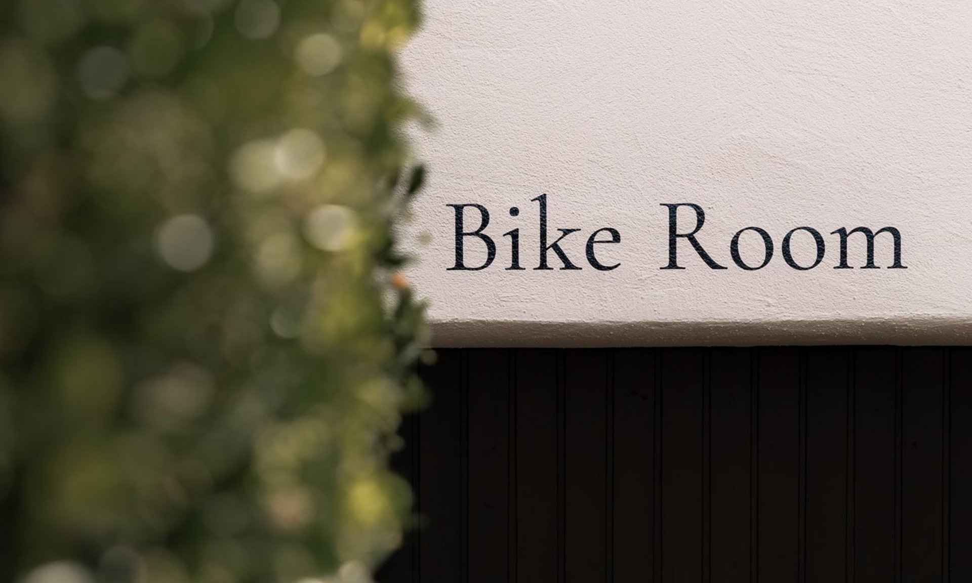 Sign reading Bike Room on white wall next to green foliage