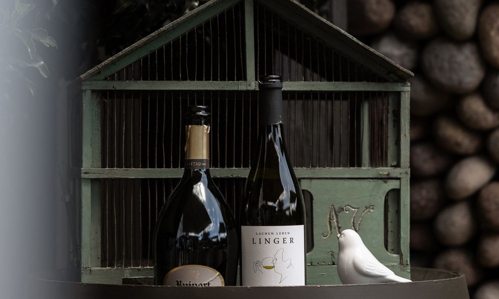 Two wine bottles on a barrel in front of a birdcage with white bird statues