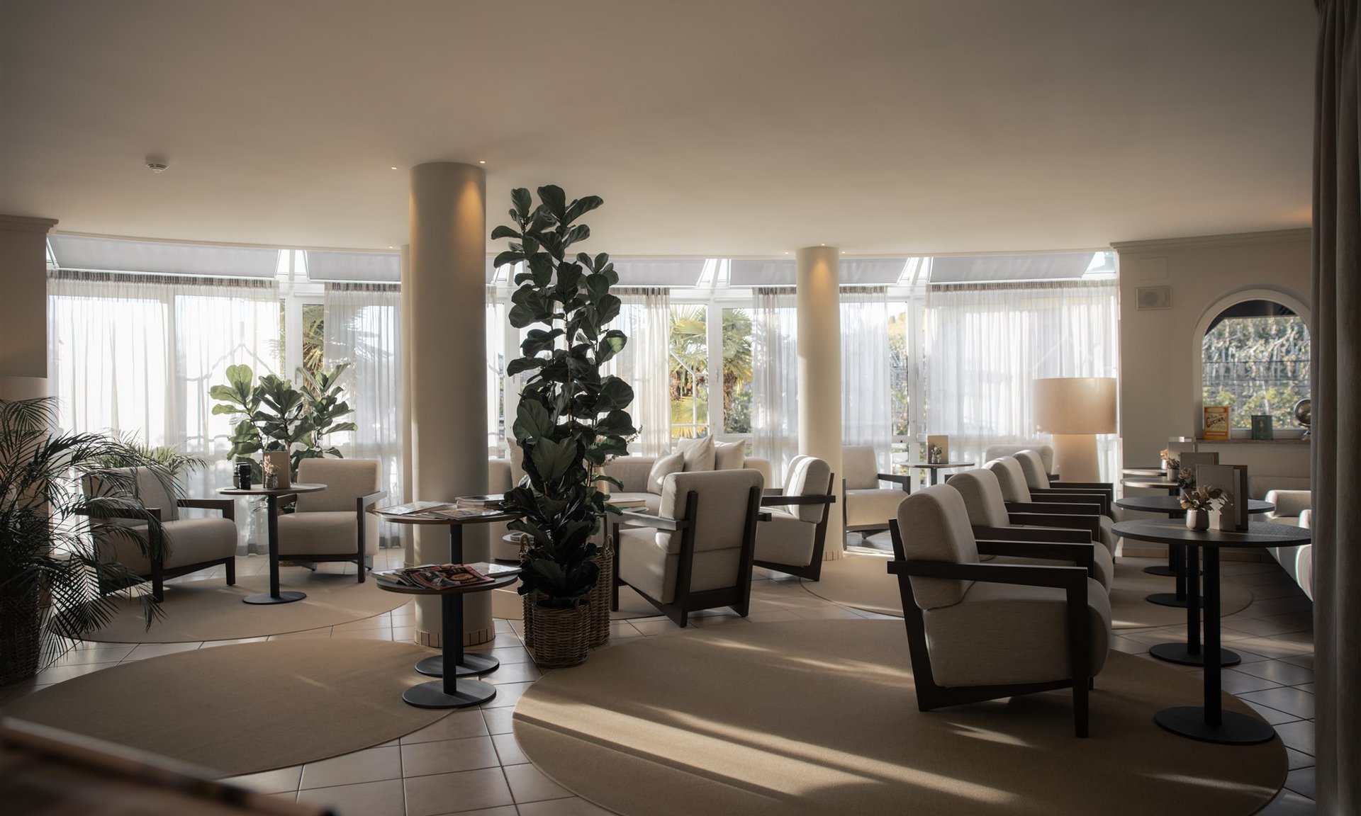 Sala d'attesa luminosa con poltrone, tavoli e piante davanti a grandi finestre