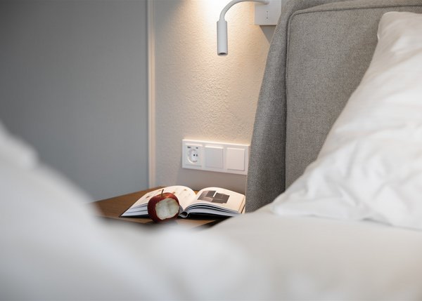 Luxus Apartment II Beleuchteter Nachttisch mit aufgeklapptem Buch und angebissenem Apfel neben einem grauen Bett